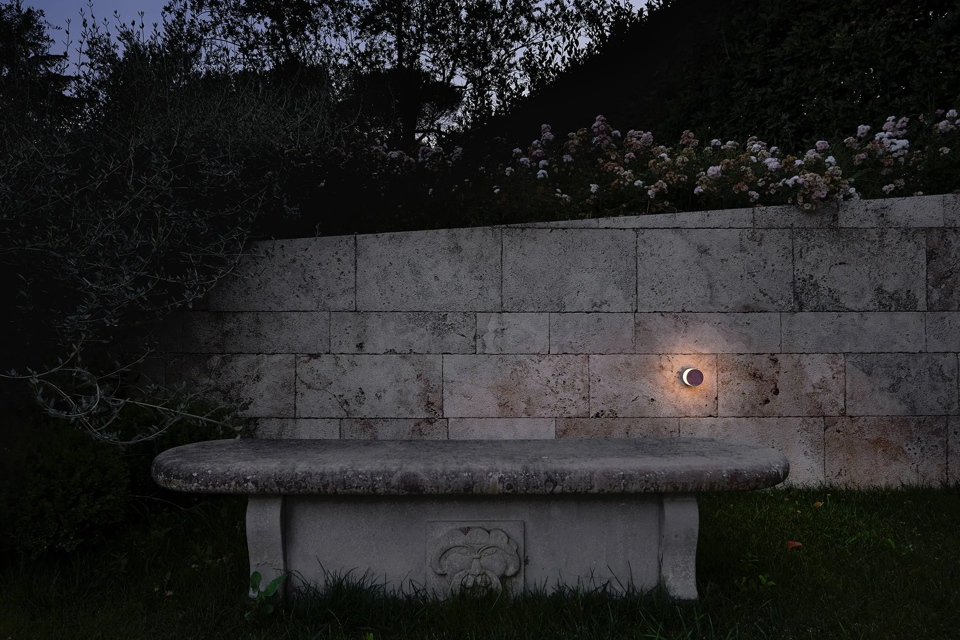 Aplique Puraluce en una pared de travertino sobre un banco de piedra en el jardín de una villa, al anochecer